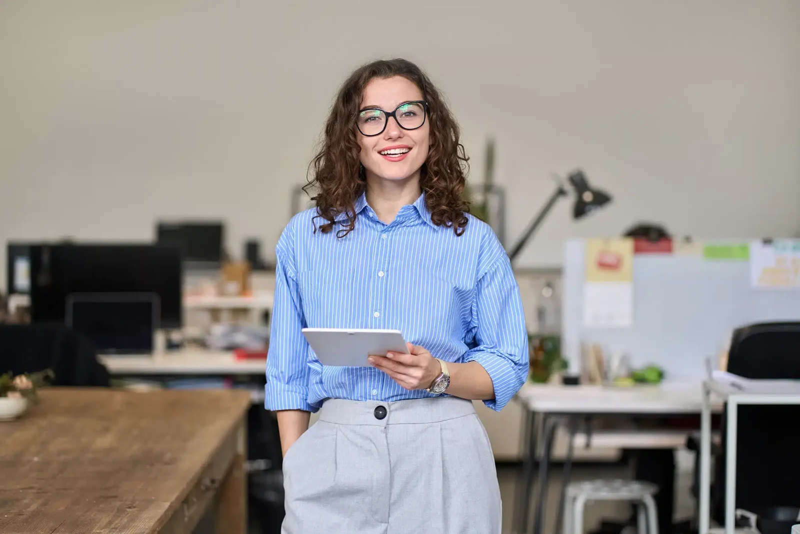 Une femme portant des lunettes et une chemise bleue tient une tablette et sourit debout dans un bureau.