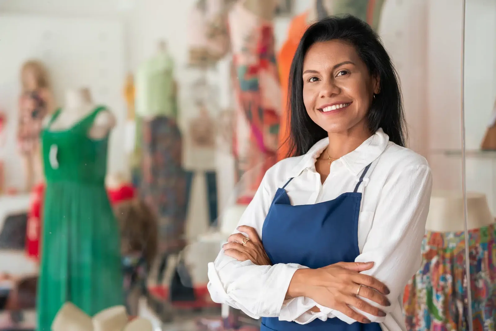 Une femme souriante aux longs cheveux noirs se tient dans un magasin de vêtements, portant une chemise blanche et un tablier bleu, les bras croisés.