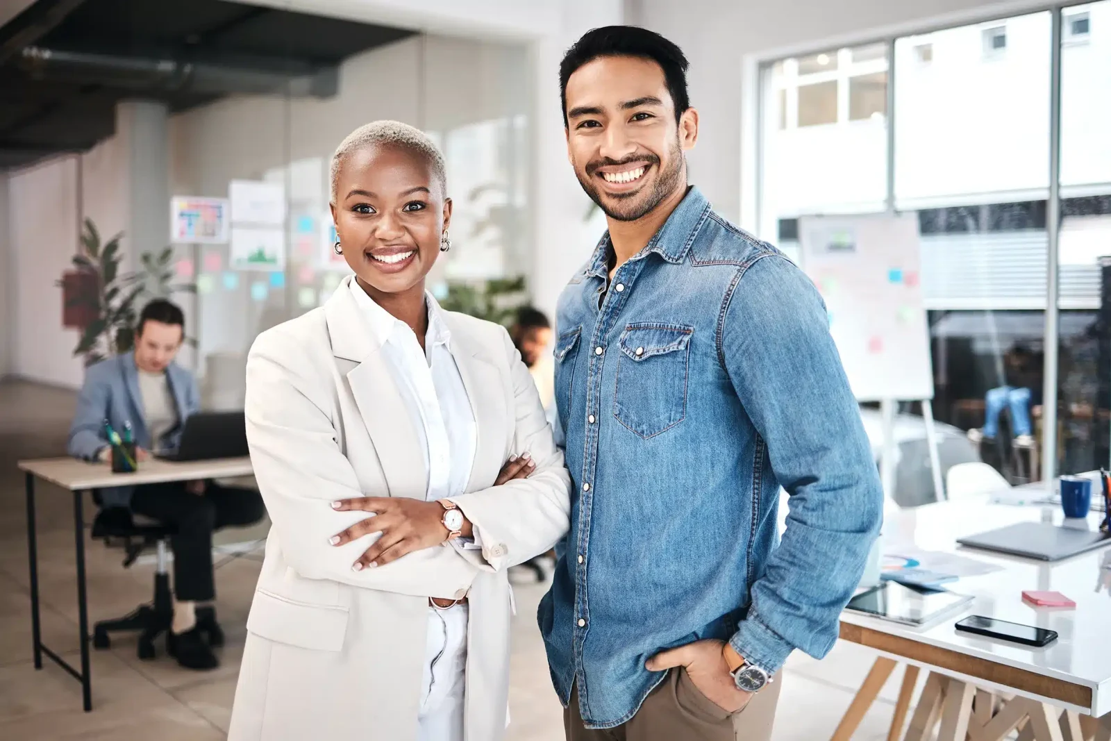 Deux individus souriants se tiennent côte à côte dans un bureau, l'un en costume blanc et l'autre en chemise en jean, avec des collègues qui travaillent derrière.