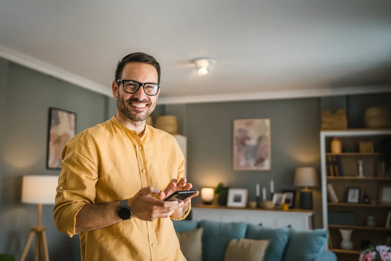 Un homme portant une chemise jaune et des lunettes sourit tout en tenant un smartphone dans un salon moderne avec des étagères et des photos encadrées.