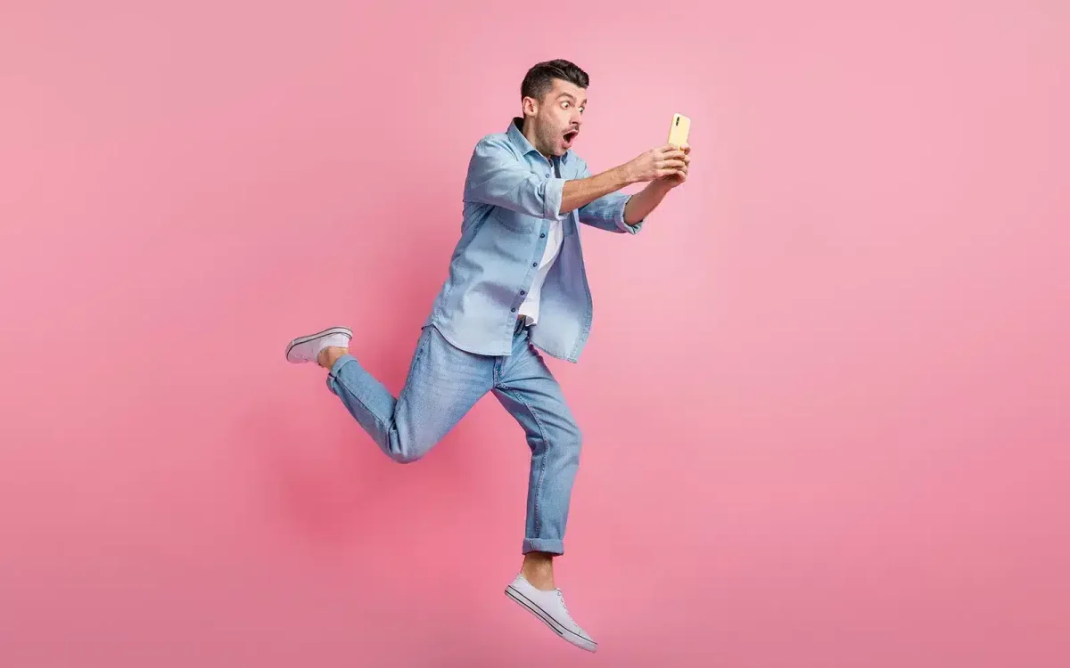 Un homme vêtu d'une tenue bleu clair saute énergiquement avec une jambe pliée et un bras tendu, prenant un selfie sur un fond rose, rappelant la façon dont vous pouvez booster l'énergie de votre site en utilisant les techniques SEO WordPress.