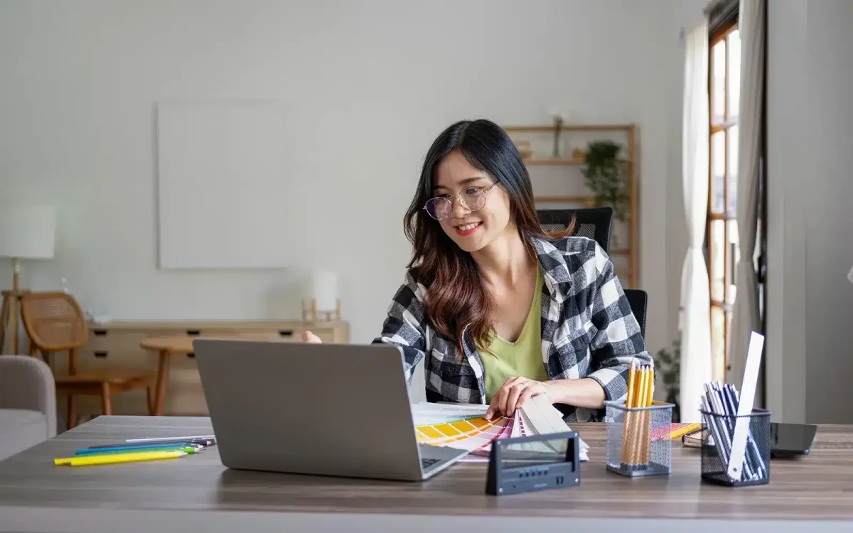 Une personne portant des lunettes sourit en regardant l'écran d'un ordinateur portable, choisissant probablement un thème WordPress. Un cahier, des échantillons de couleurs et du papier à lettres sont posés sur le bureau.