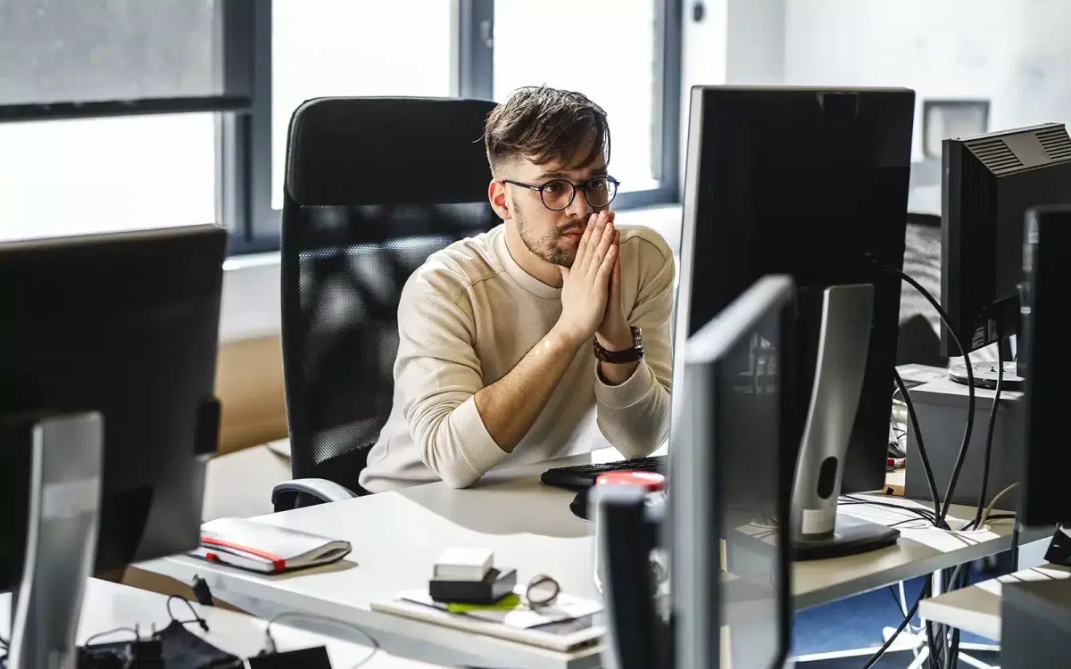 Un homme portant des lunettes est assis à un bureau avec plusieurs écrans d'ordinateur dans un bureau, regardant pensivement le moniteur, considérant les dernières extensions WooCommerce gratuites pour son site WordPress.