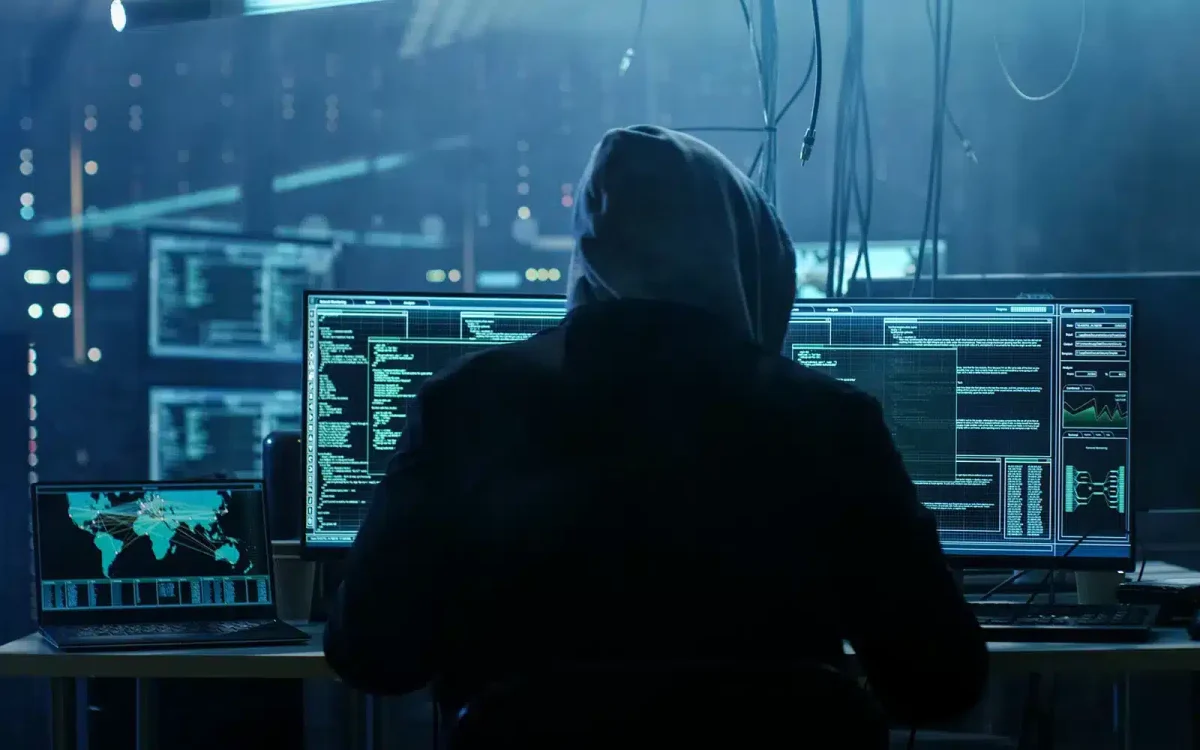 Une personne portant un sweat à capuche est assise à un bureau avec plusieurs écrans d'ordinateur affichant le code et les graphiques du site WooCommerce dans une pièce faiblement éclairée.