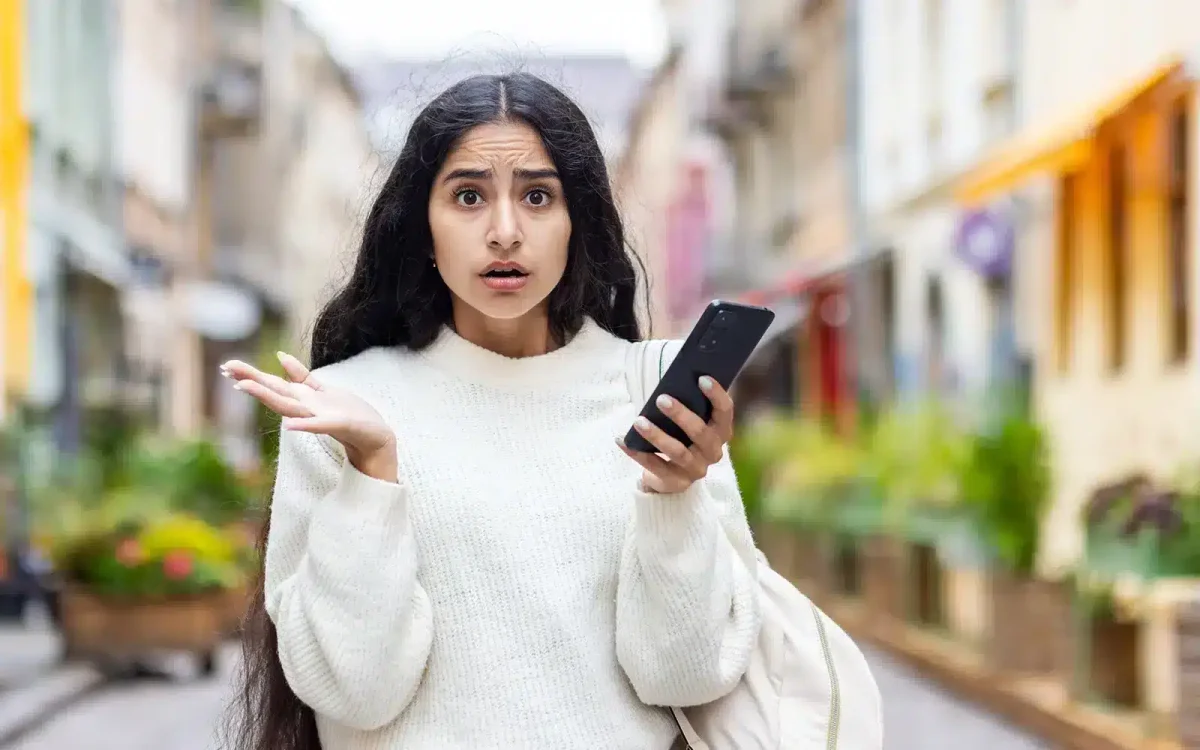 Une femme aux cheveux longs, vêtue d'un pull blanc, tient un smartphone et lève la main en signe de confusion dans une rue de la ville, comme si elle cherchait une solution au milieu de la foule animée.