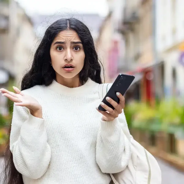Une femme aux cheveux longs, vêtue d'un pull blanc, tient un smartphone et lève la main en signe de confusion dans une rue de la ville, comme si elle cherchait une solution au milieu de la foule animée.