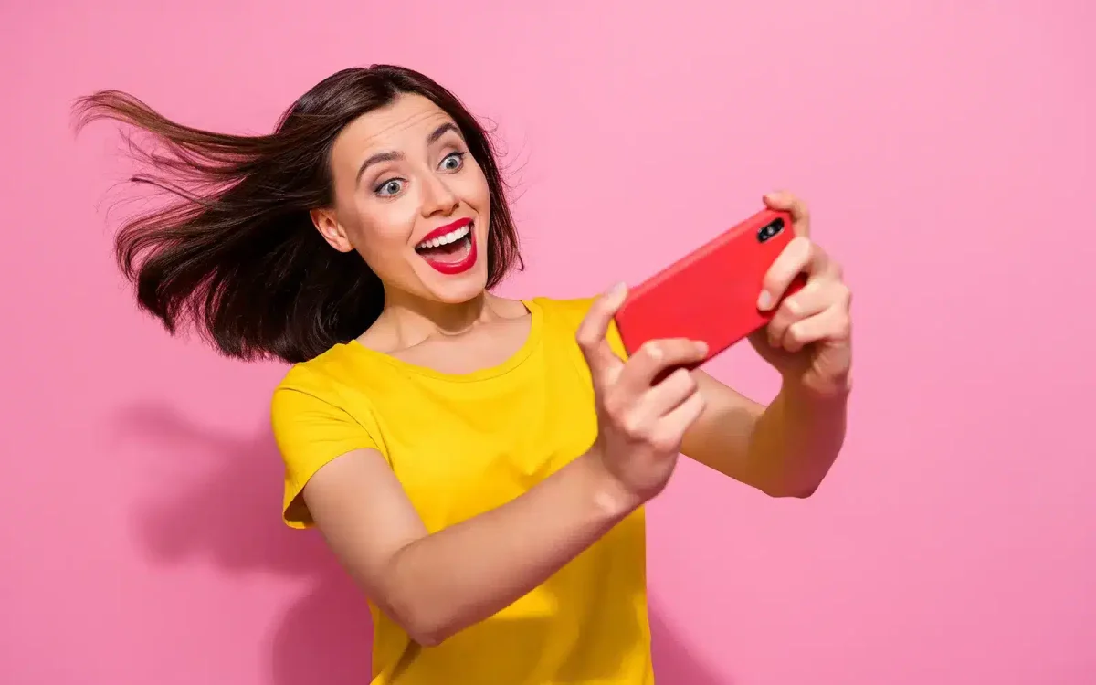 Une femme en chemise jaune prend un selfie avec un smartphone rouge sur fond rose, les cheveux flottants et l'expression excitée.
