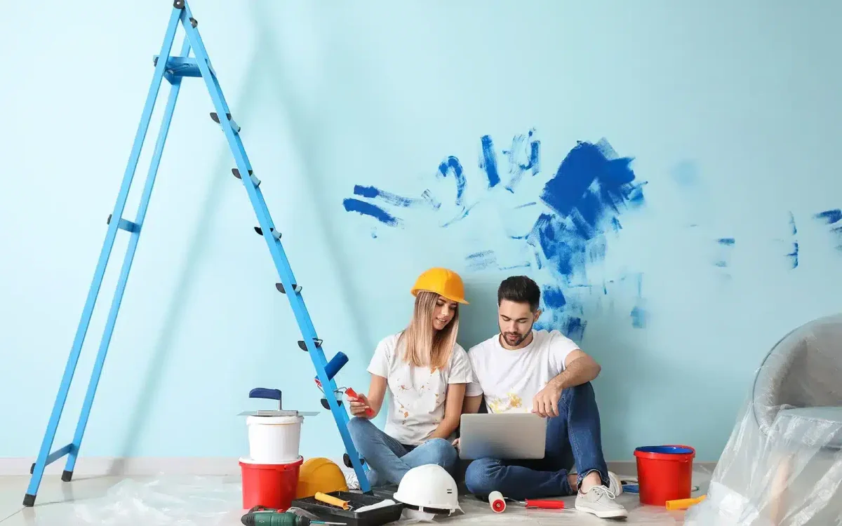 Deux personnes sont assises par terre, entourées de fournitures et d'outils de peinture, regardant un ordinateur portable ; une échelle et un mur peint en arrière-plan.