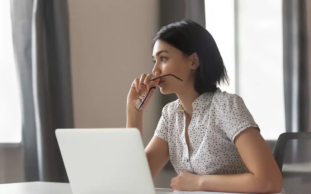 Une femme aux cheveux noirs courts portant une chemise à pois est assise à un bureau, tenant des lunettes, l'air pensif, avec un ordinateur portable devant elle.