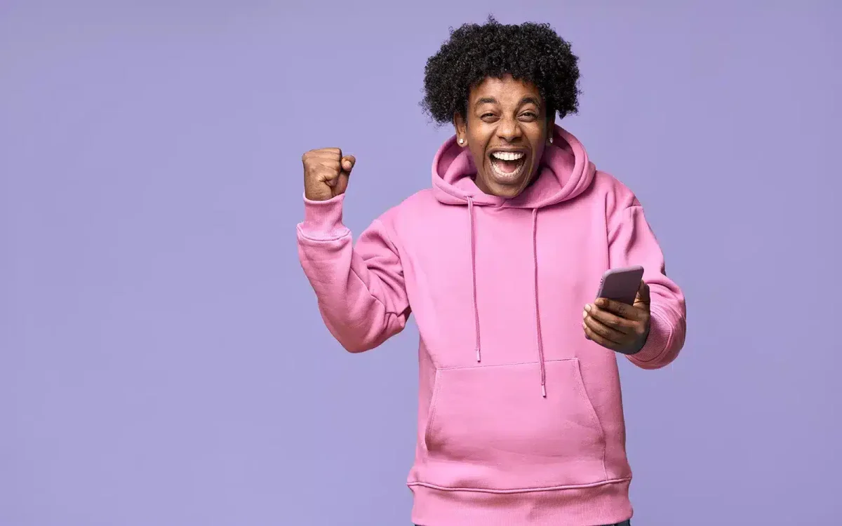 Personne portant un sweat à capuche rose souriant et levant le poing avec excitation, tenant un smartphone sur fond violet, célébrant un moment réussi de refonte de son site Web.