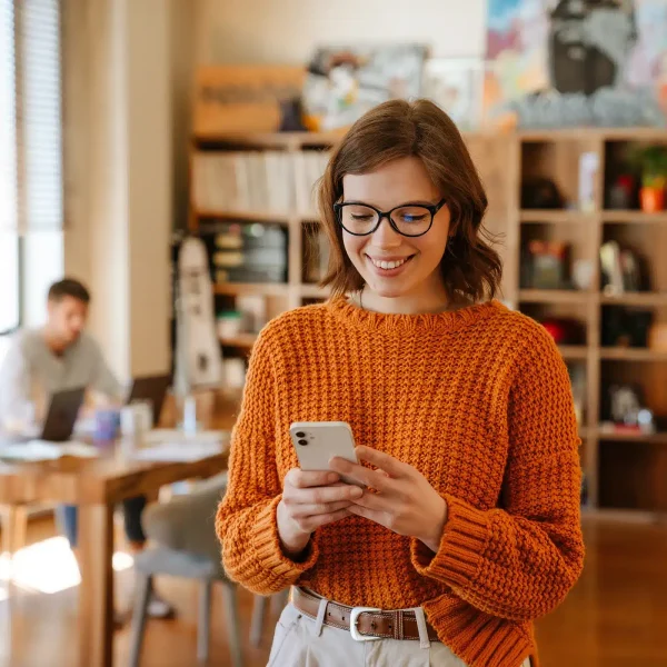 Une femme portant des lunettes et un pull orange sourit en regardant son smartphone dans une pièce très éclairée, créant un accueil chaleureux alors qu'un homme se tient en arrière-plan.