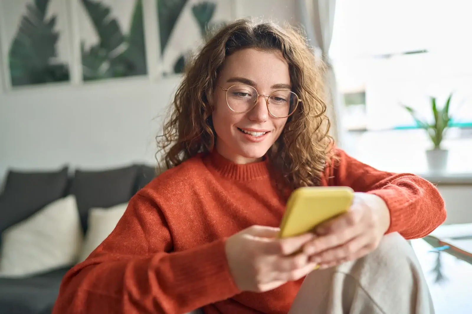 Une personne portant des lunettes et un pull rouge est assise sur un canapé, tenant et regardant un téléphone jaune tout en souriant, incarnant un accueil chaleureux.