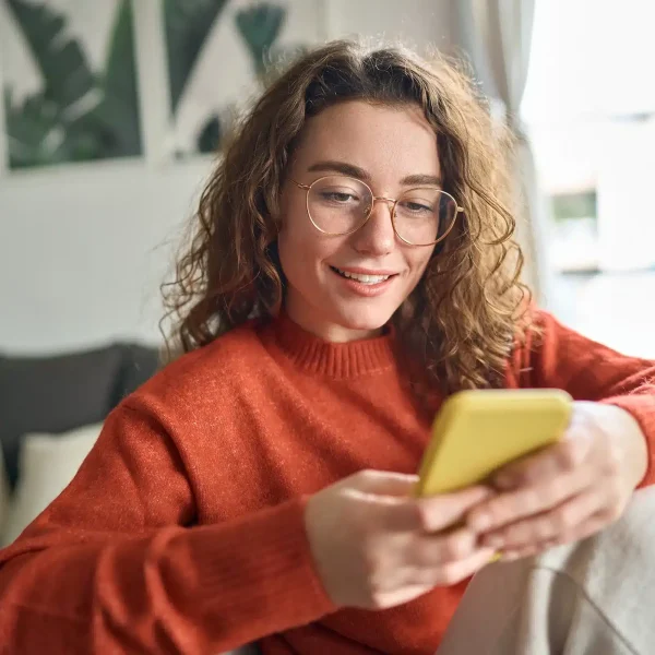 Une personne portant des lunettes et un pull rouge est assise sur un canapé, tenant et regardant un téléphone jaune tout en souriant, incarnant un accueil chaleureux.