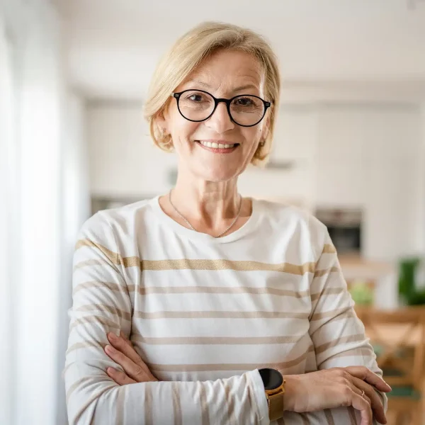 Une femme âgée portant des lunettes et des cheveux blonds courts, vêtue d'une chemise rayée, sourit et se tient debout, les bras croisés, dans un salon lumineux et accueillant.