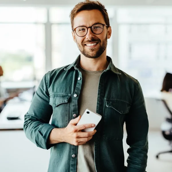 Un homme portant des lunettes et une chemise verte tient un smartphone, souriant chaleureusement dans un bureau à thème d'accueil avec des collègues flous s'affairant en arrière-plan.