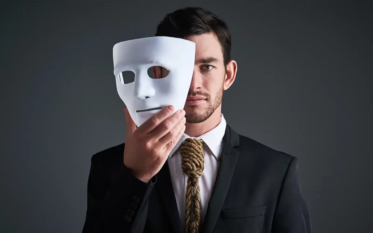 Un homme en costume avec une corde autour du cou tient un masque blanc révélant la moitié de son visage, sur un fond sombre, rappelant la façon dont l'utilisation de thèmes annulés peut masquer une véritable identité en ligne.