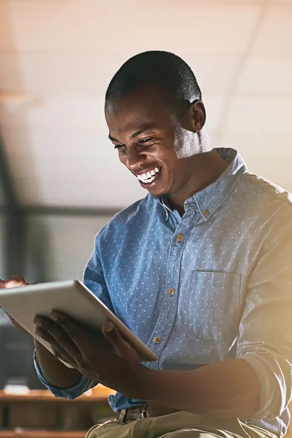 Un homme en chemise bleue sourit et utilise une tablette dans un espace de bureau faiblement éclairé, apparemment plongé dans ses pensées à propos de son projet actuel.