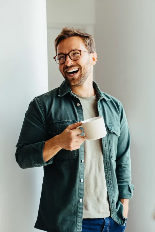 Un homme portant des lunettes et une chemise verte, tenant une tasse et souriant tout en s'appuyant contre un mur blanc, présente une introduction invitante.