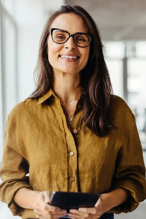 Une femme portant des lunettes et une chemise marron sourit tout en tenant une tablette dans un bureau avec des collègues en arrière-plan, discutant d'À propos.