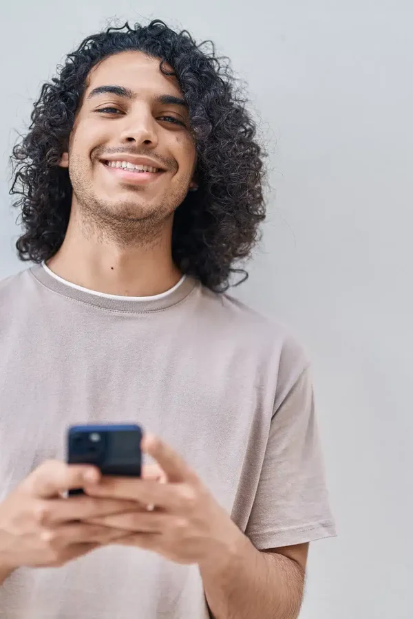 À propos, une personne aux cheveux bouclés sourit tout en tenant un smartphone sur un fond blanc uni.