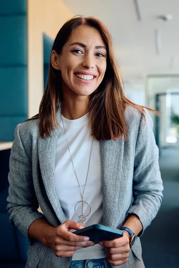 Femme souriante tenant un smartphone, debout dans un espace de bureau moderne avec des bureaux et des plantes en arrière-plan. En matière de productivité, elle incarne l'équilibre entre travail et joie.
