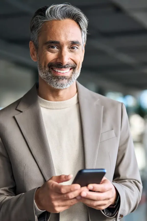 Un homme aux cheveux gris et à la barbe, vêtu d'un blazer marron, sourit tout en tenant un smartphone à l'extérieur, partageant apparemment quelque chose à propos.