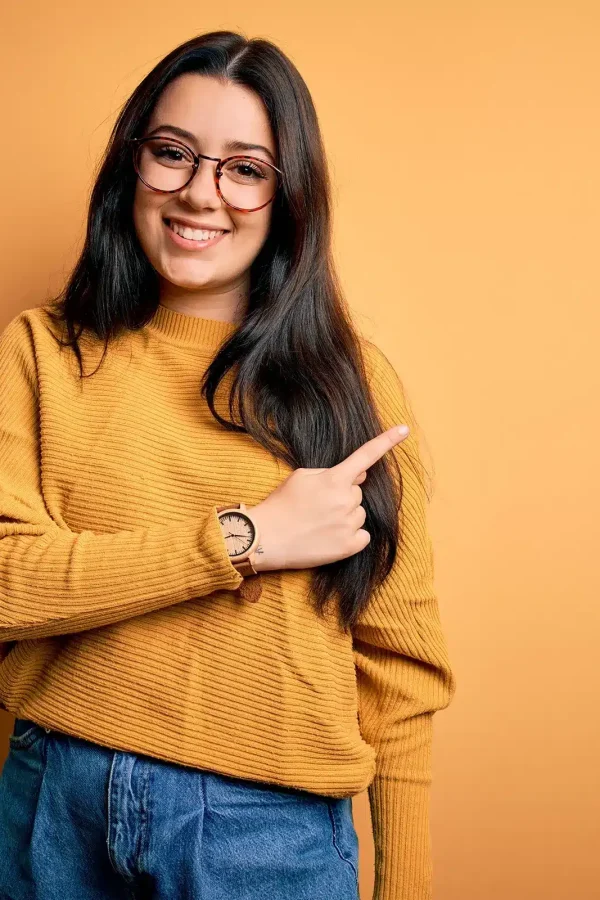Une personne souriante aux cheveux longs et noirs, portant des lunettes et un pull moutarde, pointe vers la droite sur un fond moutarde. A propos de cette scène, tout est parfaitement coordonné.