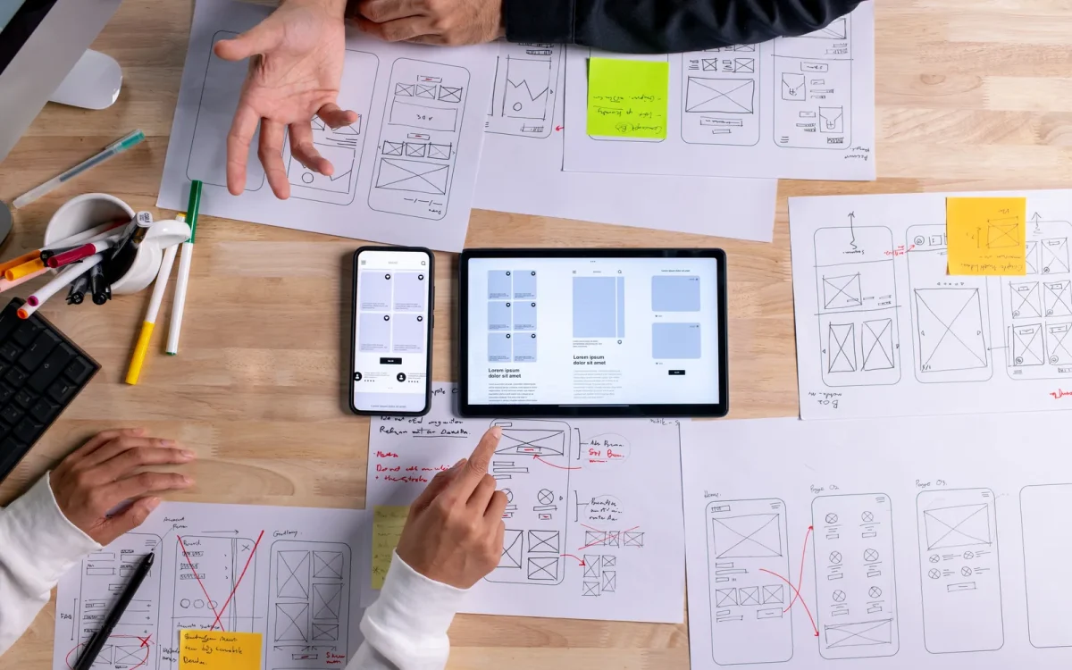 Vue de dessus de deux personnes qui annotent des maquettes de pages web sur papier, autour d'une tablette et d'un smartphone, pour illustrer un éditeur Elementor qui n'enregistre plus les modifications