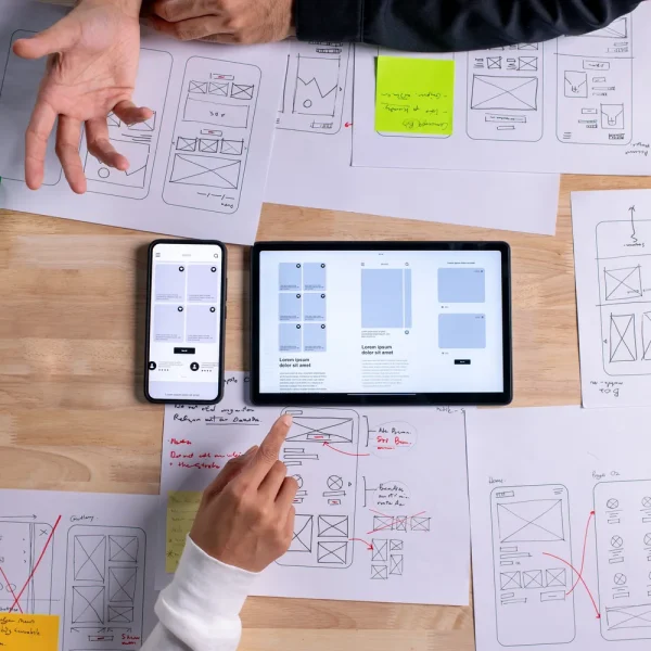 Vue de dessus de deux personnes qui annotent des maquettes de pages web sur papier, autour d'une tablette et d'un smartphone, pour illustrer un éditeur Elementor qui n'enregistre plus les modifications