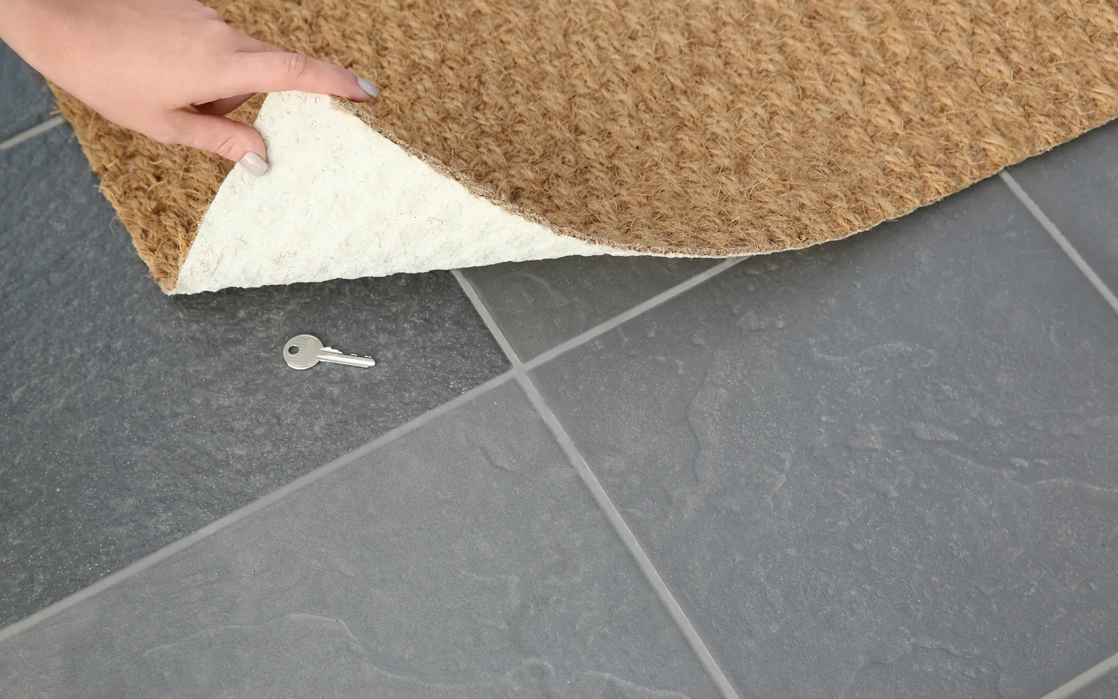 Main qui soulève un paillasson en fibre de coco et découvre une clé cachée sur le carrelage, pour illustrer les administrateurs cachés créés via la faille Ninja Forms