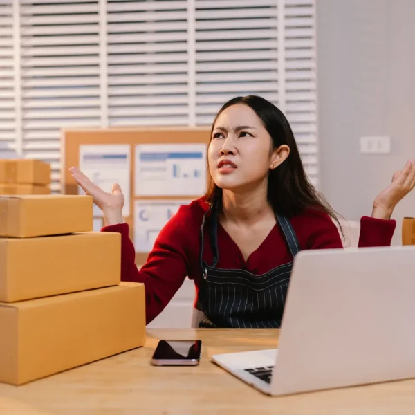 Commerçante agacée entre ses colis devant son ordinateur portable, le panier de sa boutique en ligne ne fonctionne plus
