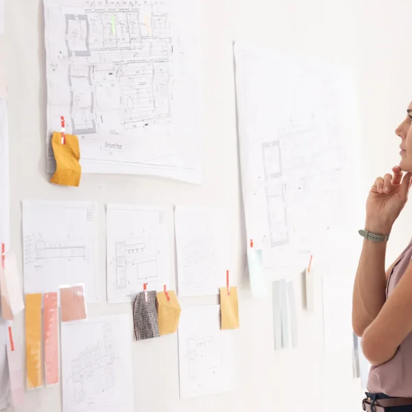 Femme pensive devant un mur couvert de plans et d’échantillons épinglés, qui prépare un projet, pour illustrer la préparation d’un site à WordPress 7.2